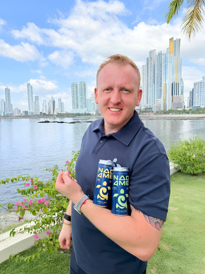 Smiling man holding NAD4ME energy drinks outdoors with city skyline and water background
